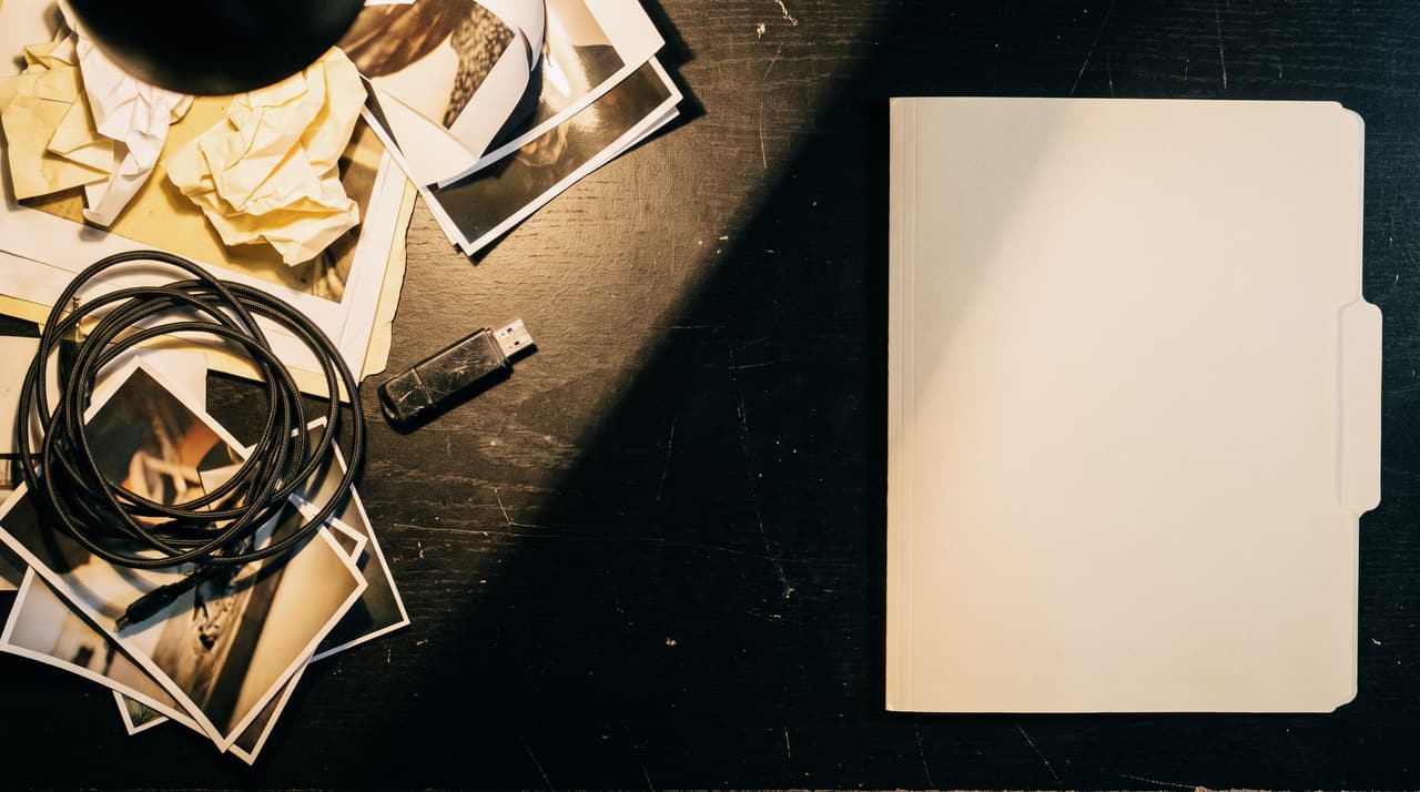 A production desk under one work light: a messy pile of paper, loose photos, a tangled cable and a USB stick on the left, one clean document folder squared to the desk edge on the right